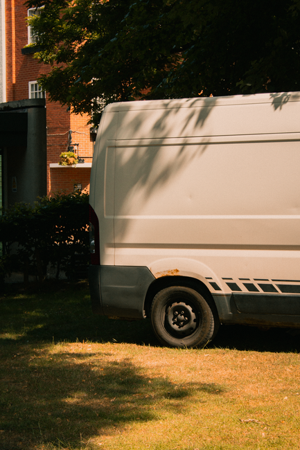 A white commercial van parked on a grassy area beside a pavement, with the rear partially visible. The van is situated next to a residential building with brick walls and windows, and is surrounded by greenery, including trees and bushes. The back door of the van is closed, and the vehicle shows signs of rust near the bottom edge. Shadows from the trees cast onto the van, indicating sunny weather. This image captures the loading area where furniture and boxes might be transferred inside for a home relocation, representing the logistics involved in house removals. The scene suggests a professional moving process, with the van prepared for loading or unloading items such as cardboard boxes, wrapped furniture, or small appliances, in connection with services like those offered by Man with Van North Woolwich during a moving or packing operation.