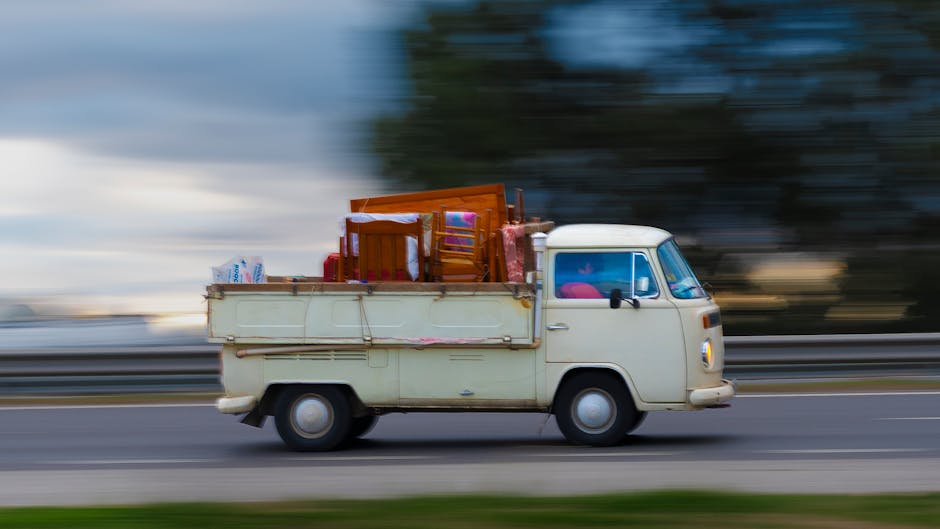 A light cream vintage van is moving along a road, with a blurred background indicating motion. The van's cargo area is loaded with various household items, including wooden chairs, a glass table, and cardboard boxes, some wrapped in plastic for protection. The furniture and boxes are arranged inside the open flatbed of the van, which is parked on the pavement of a residential area. The scene captures the loading process during a home relocation or furniture transport service, with the van operated by Man with Van North Woolwich, specialising in removals and moving logistics. The lighting suggests daytime, and the background reveals trees and sky. The focus is on the van and its contents, illustrating a typical stage in packing and moving activities associated with house removals.