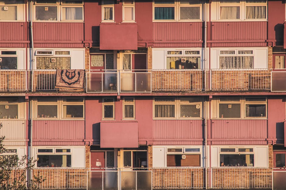 A multi-storey residential building with pink exterior walls and several balconies, each featuring metal railings and glass panels. Some balconies have personal items such as a blanket draped over the railing. Inside the building, windows with curtains or blinds are visible, with one window showing a domestic scene. The lower level displays an entrance door and multiple windows, with the shadow of the building cast onto the façade. A tree partially obscures the bottom left corner of the image. The photograph appears to be taken during daylight, with soft natural lighting illuminating the building's façade. This image relates to house removals and moving logistics, illustrating the exterior environment relevant to residential relocation services, as provided by Man with Van North Woolwich.