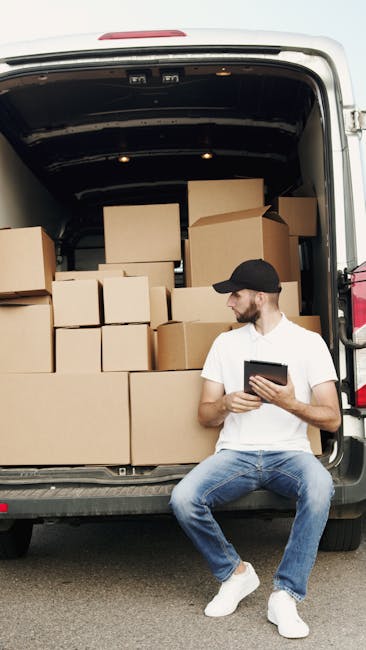 A man with a beard and wearing a white t-shirt, jeans, and a black cap sits on the pavement in front of an open van loaded with numerous cardboard boxes in various sizes. The boxes are stacked inside 