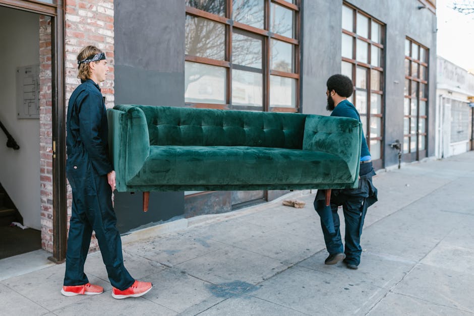 Two movers from Man with Van North Woolwich are seen participating in a home relocation, carrying a large, green upholstered sofa outside a building. One mover, wearing black pants, a blue shirt, and black shoes, holds the sofa from one side, while the other, dressed in black pants, a black jacket, and red sneakers, supports the opposite end. The sofa, with visible wooden legs, is being carefully transported through a doorway onto the pavement. The building has large windows with grid-patterned wooden framing, and the surrounding environment includes a concrete sidewalk and adjacent structures. This image illustrates the furniture transport stage of packing and moving services involved in house removals, highlighting professional handling of bulky items during the loading process outside the property.