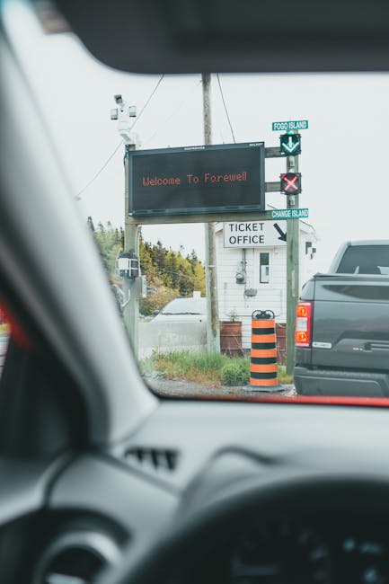 Inside a moving vehicle facing a roadside digital sign with the message 'Welcome To Farewell' in red text, positioned above a small building labeled 'Ticket Office'. The vehicle's dashboard and windshield frame the view, with other parked cars visible along the roadside. An orange and black striped traffic bollard is situated next to the building, and a utility pole with wires extends upward behind the sign. The scene depicts a moment during a home relocation or moving process, with the digital sign indicating a farewell message, and the vehicle likely en route to or from a transfer point managed by Man with Van North Woolwich, a professional removals service specializing in home and furniture transport.