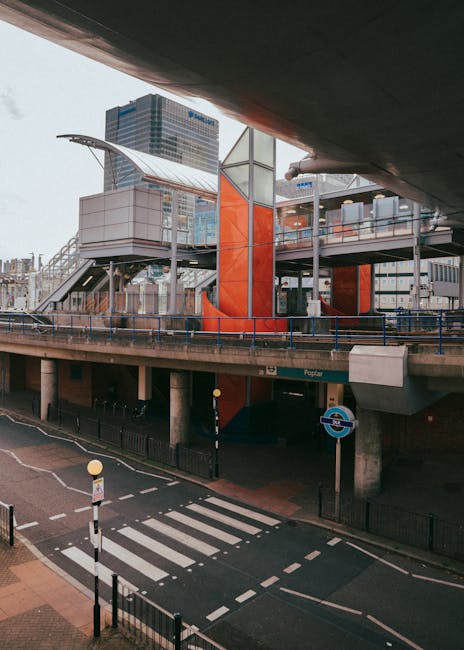 A view of an urban transportation hub with a multi-level structure featuring external stairs and glass panels, situated above a street with a pedestrian crosswalk and a roundel indicating the London Underground. The scene is captured during daylight with modern office buildings in the background. The image also shows part of a dark overhanging section of a building or bridge at the top, casting shadow over the area beneath. This setting reflects the logistical environment where house removals and furniture transport could take place, illustrating the external access points and urban infrastructure relevant to home relocation services provided by Man with Van North Woolwich, as described on their King George V DLR moving access guide for locals.