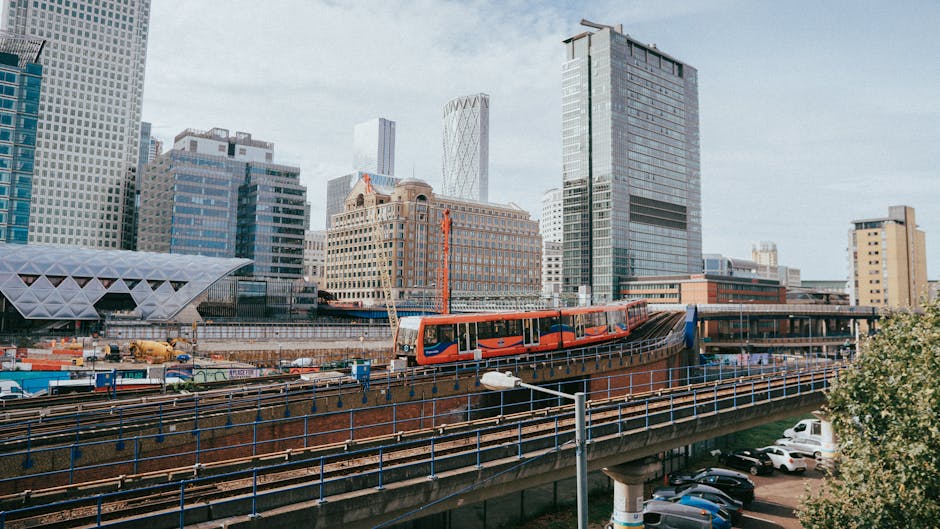 A view of an urban transportation hub with a multi-level structure featuring external stairs and glass panels, situated above a street with a pedestrian crosswalk and a roundel indicating the London Underground. The scene is captured during daylight with modern office buildings in the background. The image also shows part of a dark overhanging section of a building or bridge at the top, casting shadow over the area beneath. This setting reflects the logistical environment where house removals and furniture transport could take place, illustrating the external access points and urban infrastructure relevant to home relocation services provided by Man with Van North Woolwich, as described on their King George V DLR moving access guide for locals.