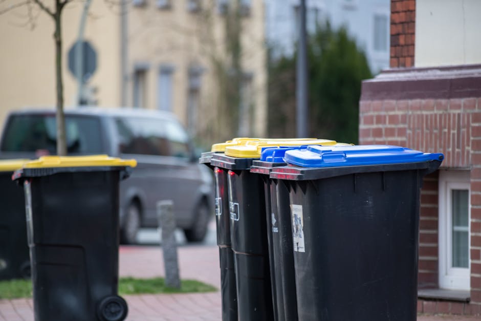 A row of four large outdoor wheelie bins with black bodies and yellow, blue, and green lids are positioned on a paved pavement near a brick building, with one bin slightly ahead of the others. Behind the bins, a parked grey van is visible on the street, and the background includes trees and residential buildings under natural daytime lighting. The scene depicts waste collection and disposal in an urban area, relevant to house removals and relocation logistics, with [COMPANY_NAME] involved in the moving or packing process near North Woolwich, as referenced in the page about Newham Council's bulky waste rules.