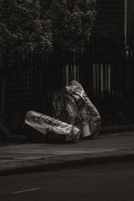 Three large black plastic garbage bags filled with household waste and debris are positioned on the pavement outside a property, leaning against a metal fence and a brick wall partially obscured by trees in the background. The bags appear to contain unknotted recyclables or discarded materials, with some cardboard packaging visible on top. The setting is during daytime with natural lighting, and the bags are situated near a street curb, likely awaiting collection. This scene relates to general waste disposal and not directly to house removals or moving services, but it aligns with waste management considerations involved in home relocation projects provided by Man with Van North Woolwich, especially under the context of complying with local bulky waste rules in Newham.