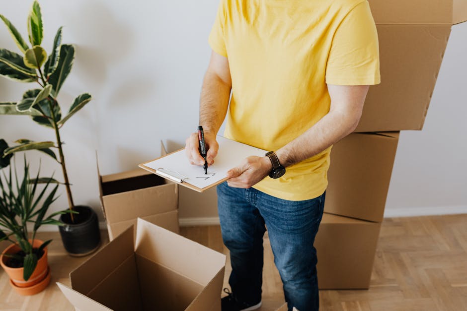 A man with dark skin, a full beard, and curly black hair wearing a blue t-shirt, is standing in a room surrounded by moving boxes made of cardboard, some with black tape seals, and a large cardboard b