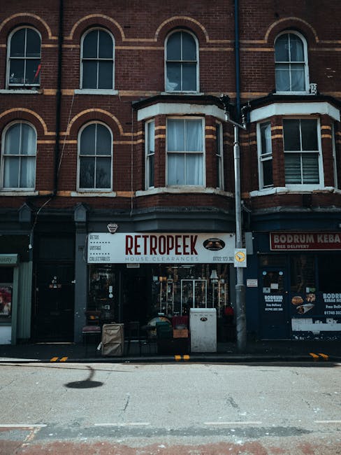 Exterior view of a multi-storey brick building on a city street, featuring large arched windows on the upper floors and ground-floor shops with signage, including a sign for RETROPEEK house clearances and another for Bodrum Kebab. In front of the shops, there are several cardboard boxes, plastic crates, and a small trolley on the pavement, indicating a moving or clearance process. The scene is captured during daytime with natural lighting, and the street appears quiet with no vehicles or pedestrians visible. This setting relates to house removals and relocation services, illustrating the process of moving items out of a property with professional assistance from [COMPANY_NAME].