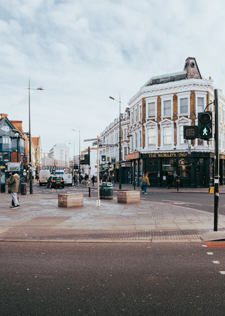 A busy street scene in North Woolwich showing a commercial pedestrian crossing with a green pedestrian light. In the foreground, there are wooden pallets placed on a paved area, likely used for loading or temporary storage during home relocation or furniture transport. To the left, a person is walking past a shop with large windows, while another person in yellow stands nearby. The street is lined with multi-storey buildings, including a corner building with a curved white façade and decorative architectural details, housing a pub or café. Vehicles, including cars and a double-decker bus, are visible further down the street, suggesting active traffic and movement typical of a retail or commercial zone. This image reflects the urban environment where moving services like those provided by Man with Van North Woolwich might facilitate packing and moving activities, with the loading process occurring in the street area under daytime lighting.