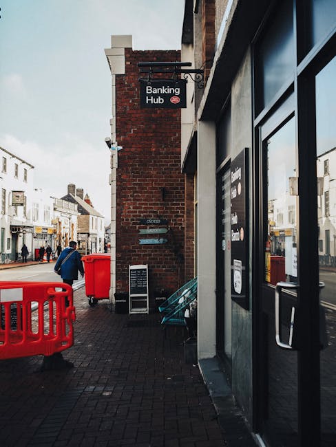 Exterior view of a multi-storey brick building on a city street, featuring large arched windows on the upper floors and ground-floor shops with signage, including a sign for RETROPEEK house clearances and another for Bodrum Kebab. In front of the shops, there are several cardboard boxes, plastic crates, and a small trolley on the pavement, indicating a moving or clearance process. The scene is captured during daytime with natural lighting, and the street appears quiet with no vehicles or pedestrians visible. This setting relates to house removals and relocation services, illustrating the process of moving items out of a property with professional assistance from [COMPANY_NAME].