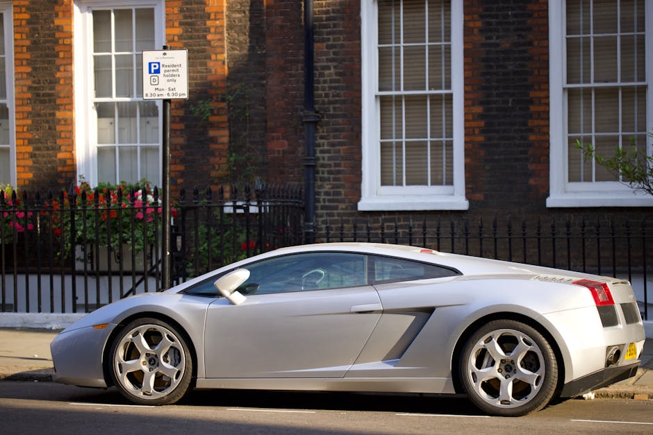 A silver two-door sports car parked on the side of a residential street in front of a brick house with white window frames. The car is positioned parallel to the curb, with its front facing left. The vehicle features a sleek, aerodynamic design with a low profile, alloy wheels, and tinted windows. Behind the car, there is a black metal fence enclosing a small garden area with a flower bed containing pink and red flowers. A parking permit sign indicating restrictions for residents only, with specified times, is mounted on a pole adjacent to the house. The scene is illuminated by natural daylight, and the surroundings include typical urban greenery and pavement, reflecting a quiet urban neighbourhood. This setting aligns with home relocation activities, involving the loading or unloading of belongings during a move conducted by Man with Van North Woolwich, with the vehicle possibly used for furniture transport or packing and moving tasks.