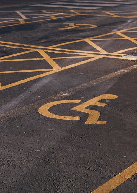 Close-up view of a section of a parking lot surface featuring yellow painted lines and wheelchair symbols, indicating designated accessible parking spaces. The asphalt appears clean with visible tire marks and slight wear around the markings. The yellow lines and symbols are clearly visible and well-maintained, highlighting the accessible parking area adjacent to likely the property involved in a home relocation. This scene represents the area where vehicles, including vans used by [COMPANY_NAME], may load or unload furniture and household items during packing and moving processes, supporting the procedures described in the Silvertown removals guide for parking permit advice for Newham, NORTH WOOLWICH. The image emphasizes parking organization essential for smooth loading and unloading operations typical of professional removals services.